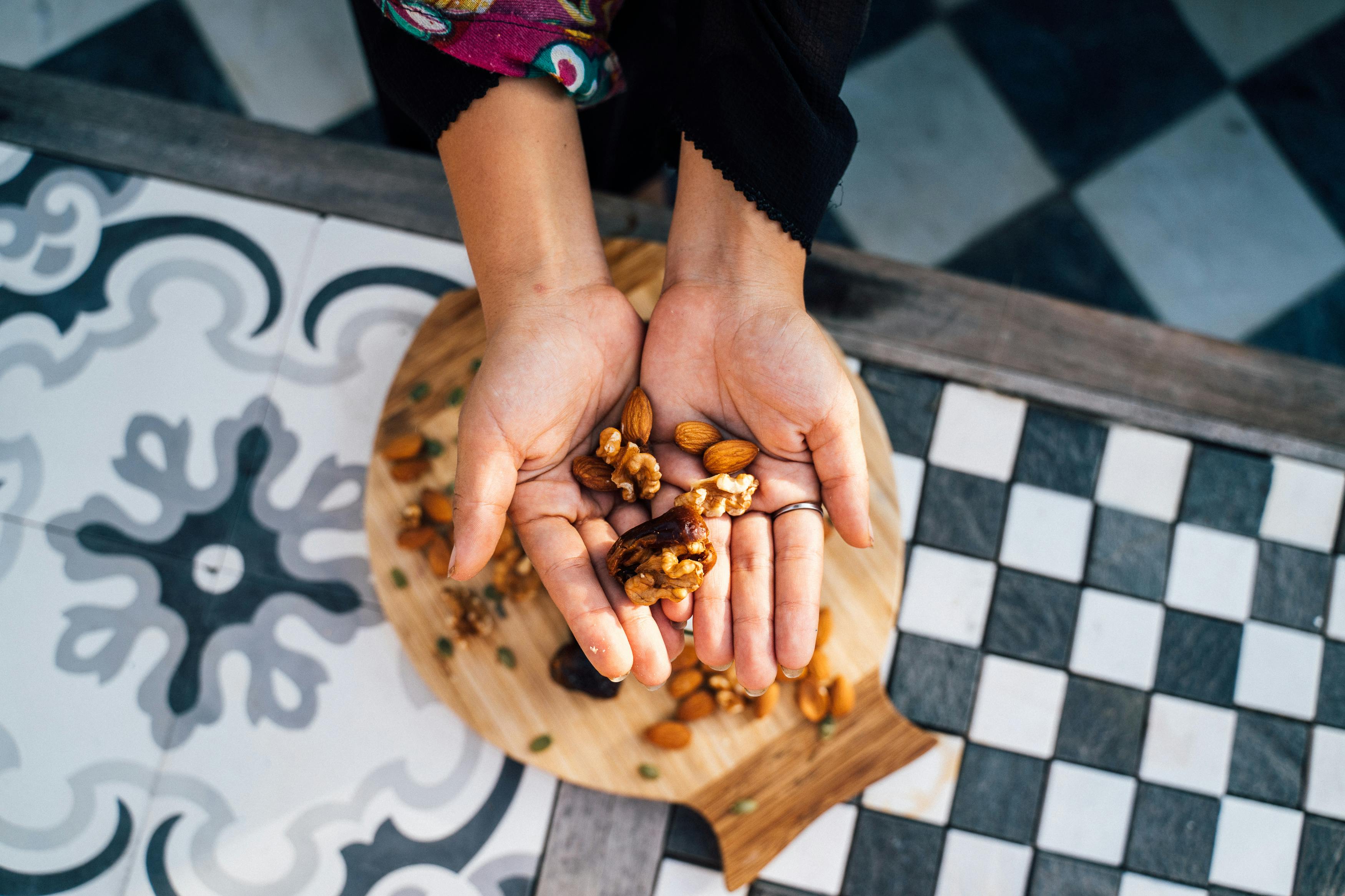 Hands hold mixed nuts over a decorative tile, showcasing almonds and walnuts in a flatlay style.